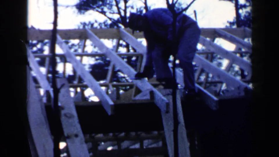 A worker bends over the exposed wooden rafters, with trees and sky visible beyond the unfinished roof frame.