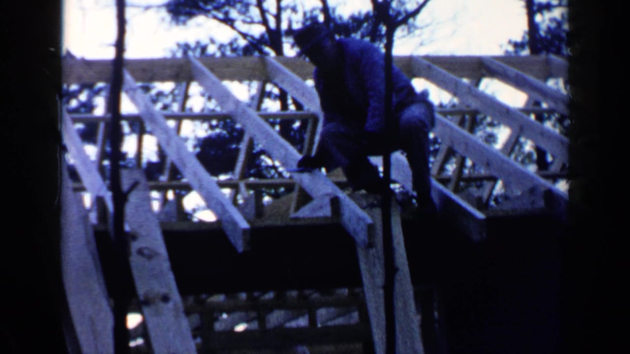 Man crouched among wooden roof joists with trees in the foreground.