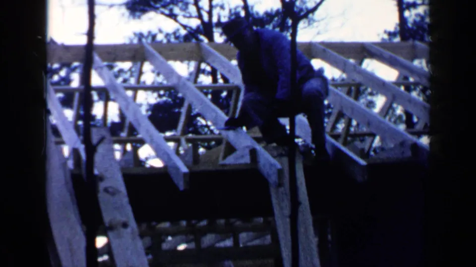 In 1957 Birmingham home-movie footage, a man works among pale wooden roof joists partly obscured by tree trunks.