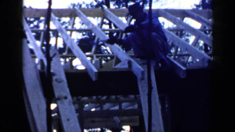 A man in dark clothing works on a wooden roof frame in Birmingham, Alabama, with trees partially blocking the 8mm view.