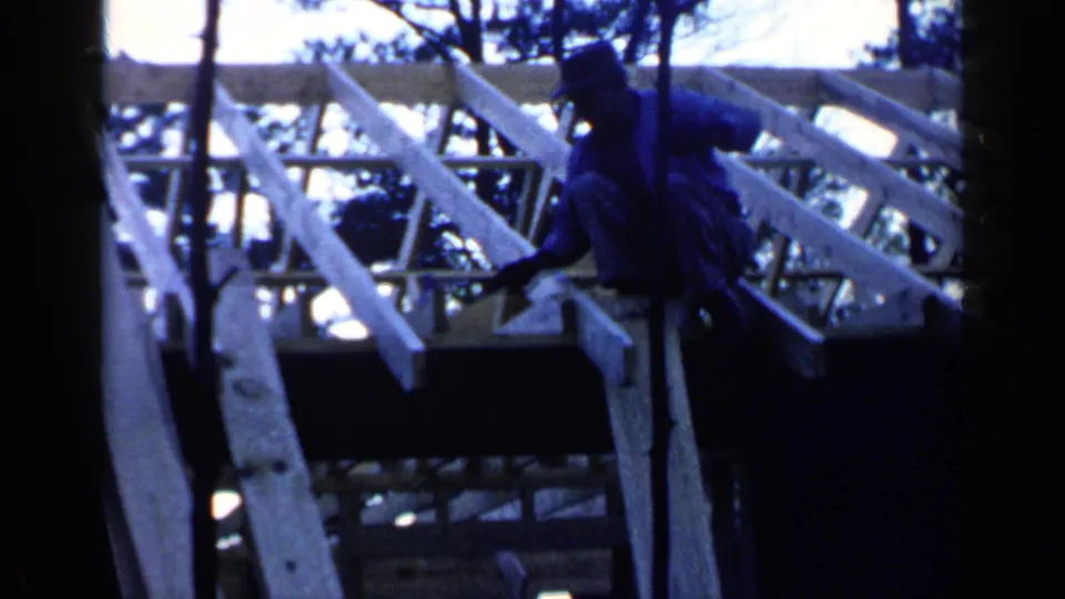 Blue-tinted 1957 8mm footage shows a man leaning over roof joists during construction work in Birmingham.