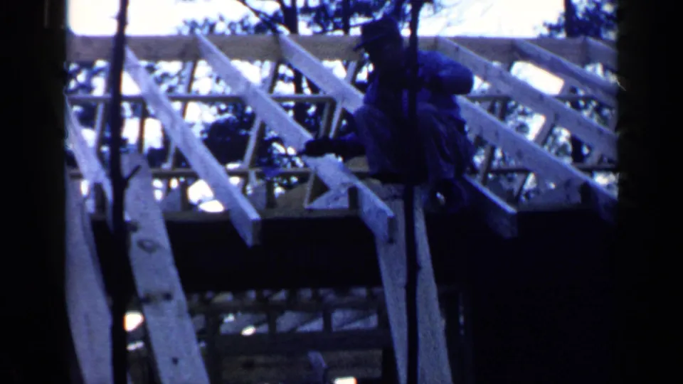The Birmingham construction scene continues as the worker crouches near exposed wooden joists, partly hidden by trees.