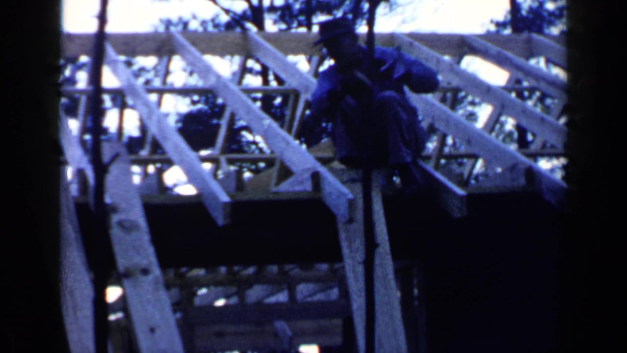 Worker in dark clothing on a wooden roof frame with rafters and trees behind him.