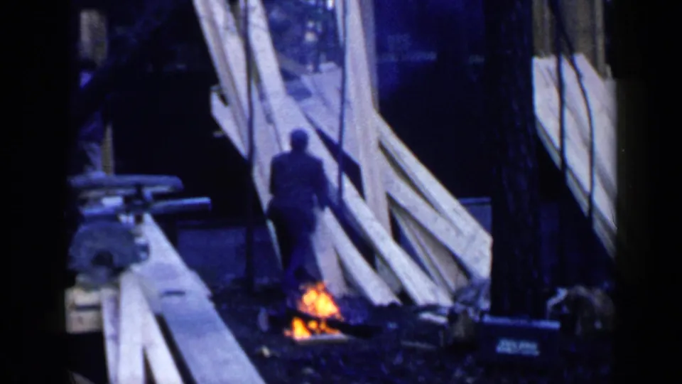 A man walks through a Birmingham construction site near stacked boards and a small ground fire, with large lumber leaning nearby.