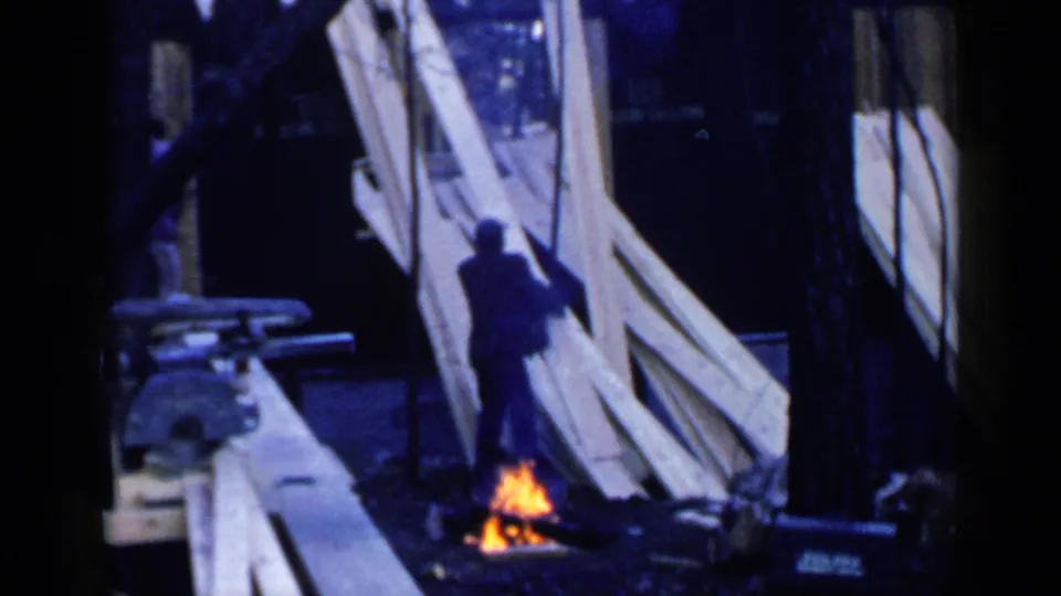 At the Birmingham worksite, a man stands beside long framing boards with a small orange fire burning near his feet.