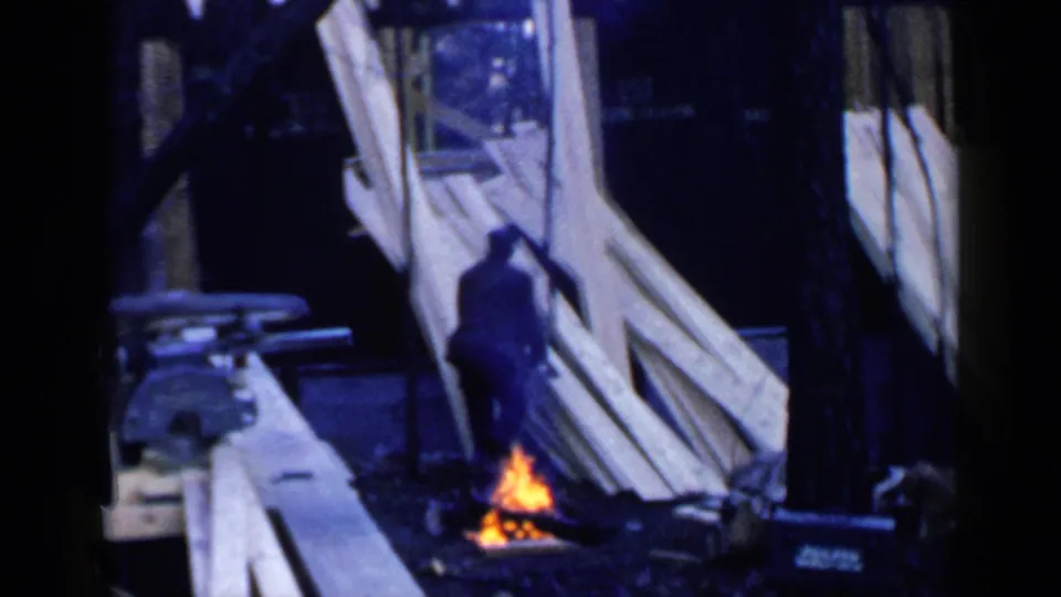 A worker bends or steps near the small fire at the 1957 Birmingham building site, surrounded by lumber and tree shadows.