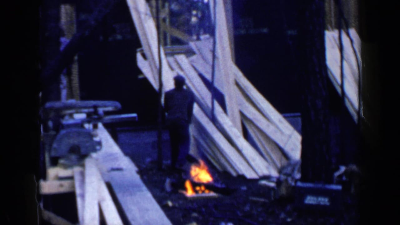 Man near stacked lumber and a small fire at an outdoor construction site.