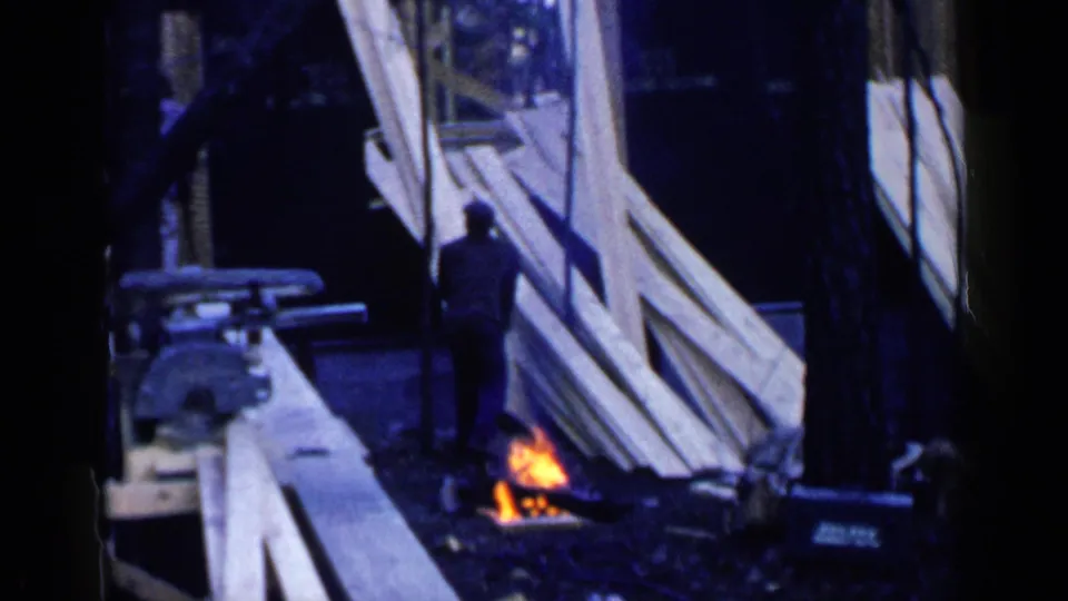1957 Birmingham home-movie frame showing a man by pale lumber boards and a small ground fire near a partially built structure.