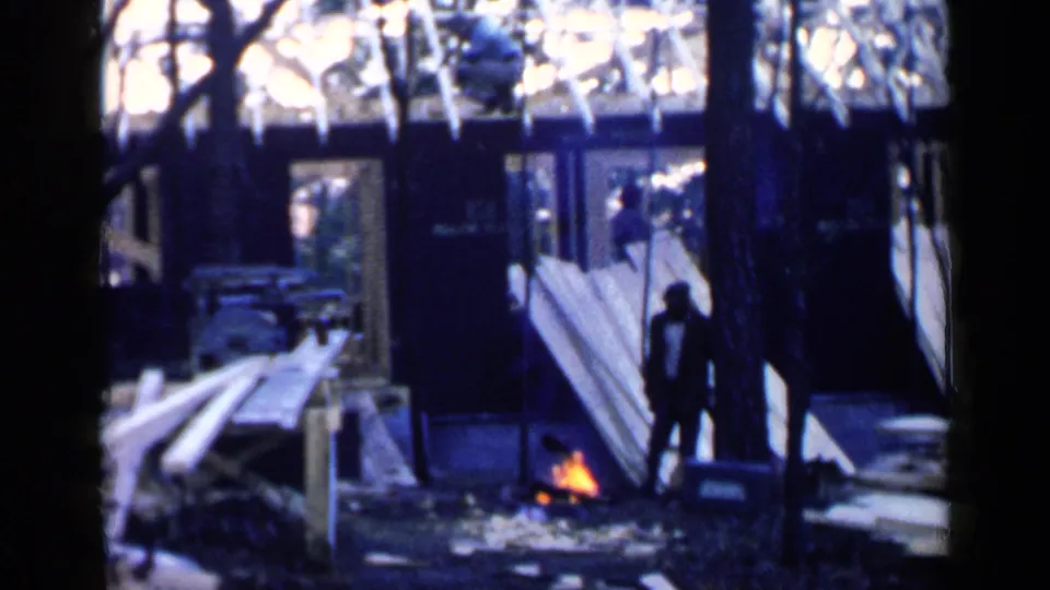 1957 Birmingham home-movie frame of a man standing near a small ground fire, with lumber piles and a partially framed structure behind him.