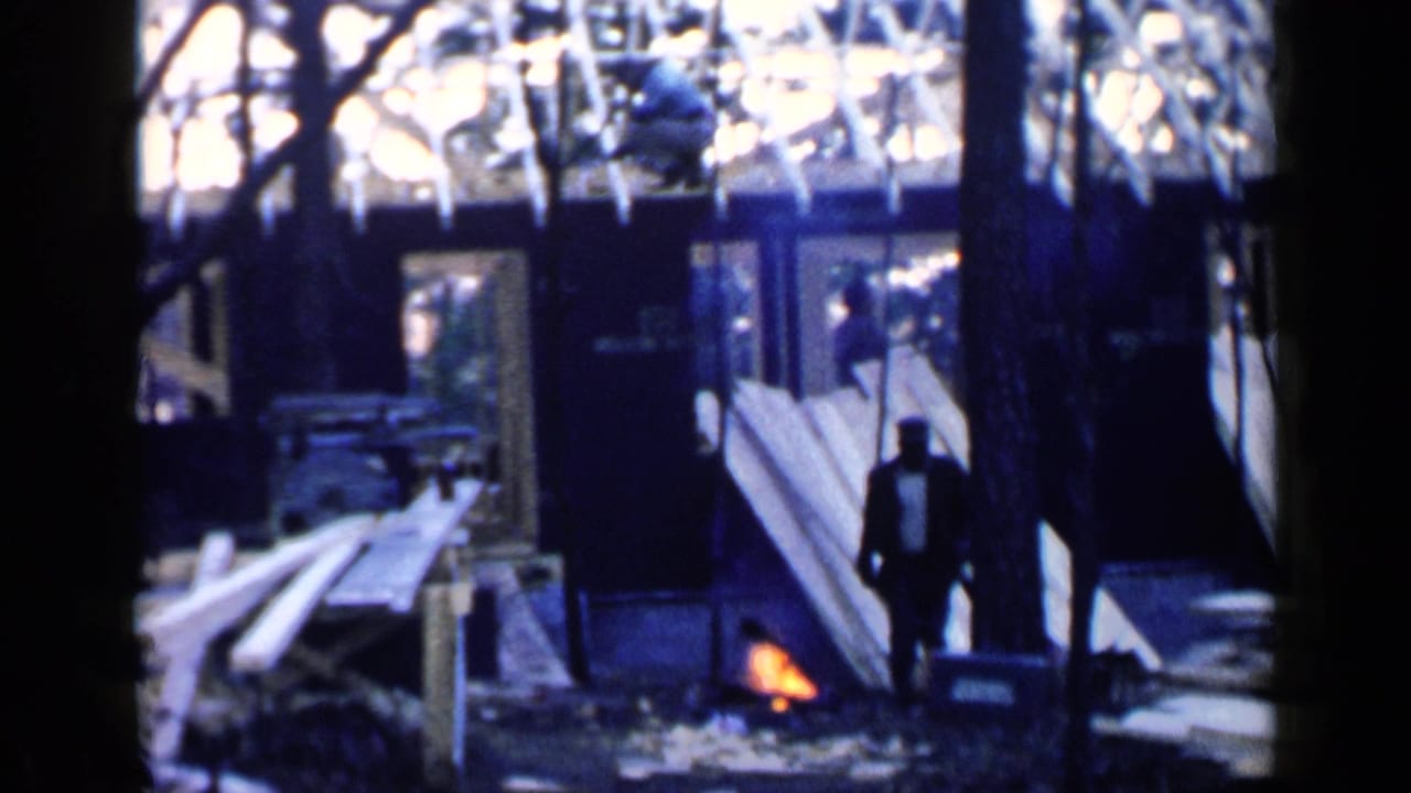 Small campfire and silhouetted figure near boards outside a dark wooden structure.