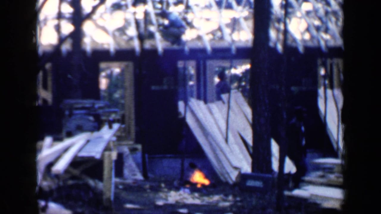 Campfire burns beside lumber while a figure stands near a wooded structure.