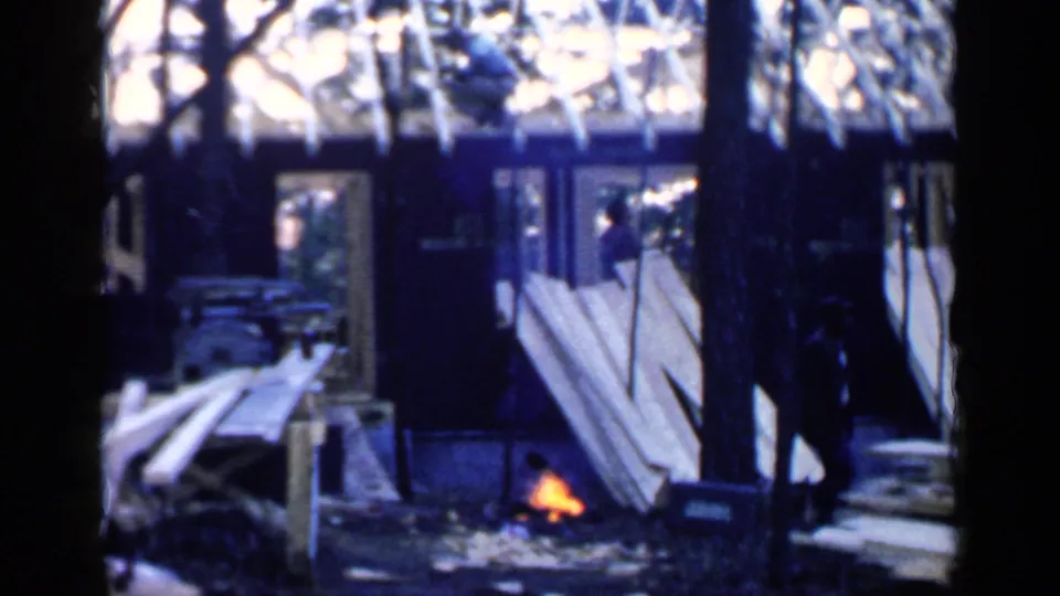 A 1957 Birmingham home-movie frame shows a fire beside piled boards, with a silhouetted person near a dark structure among trees.