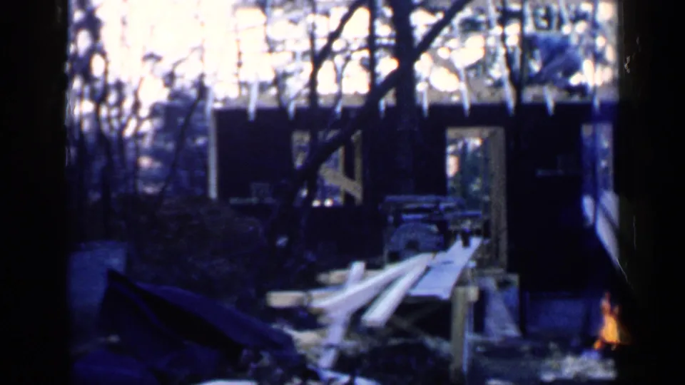 1957 Birmingham 8mm frame with stacked planks, a dark doorway, bare trees, and a small fire partly visible at the edge.