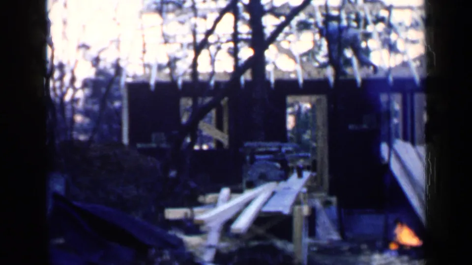 A 1957 Birmingham home-movie frame shows a rustic wooded site with a dark structure, boards, trees, and a small campfire.