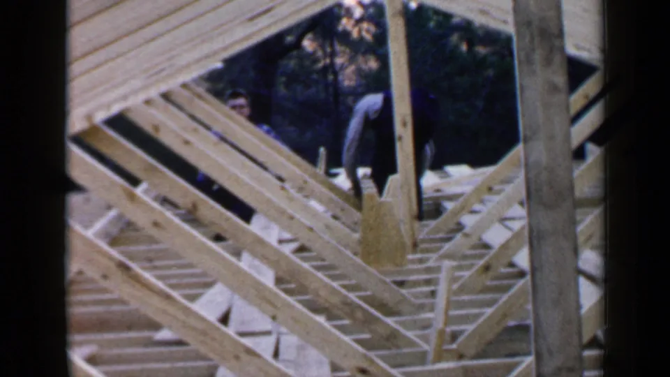 A man appears on a wooden house frame in Birmingham, Alabama, with diagonal framing, joists, vertical posts, and trees behind the structure.