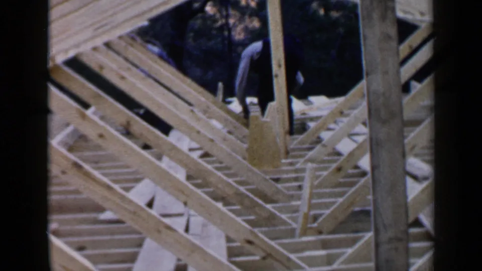 Birmingham 1957 home-movie frame showing exposed lumber framing, repeated joists, a vertical post, and a partially visible man near the top of the structure.