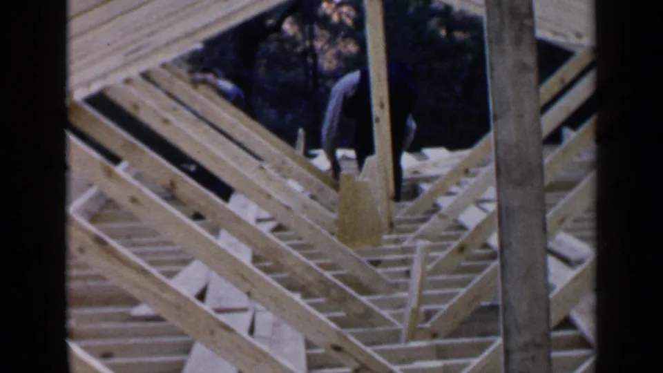 A 1957 Birmingham home-movie view of a house frame, showing open joists, diagonal braces, a vertical post, and a worker partly visible near trees.