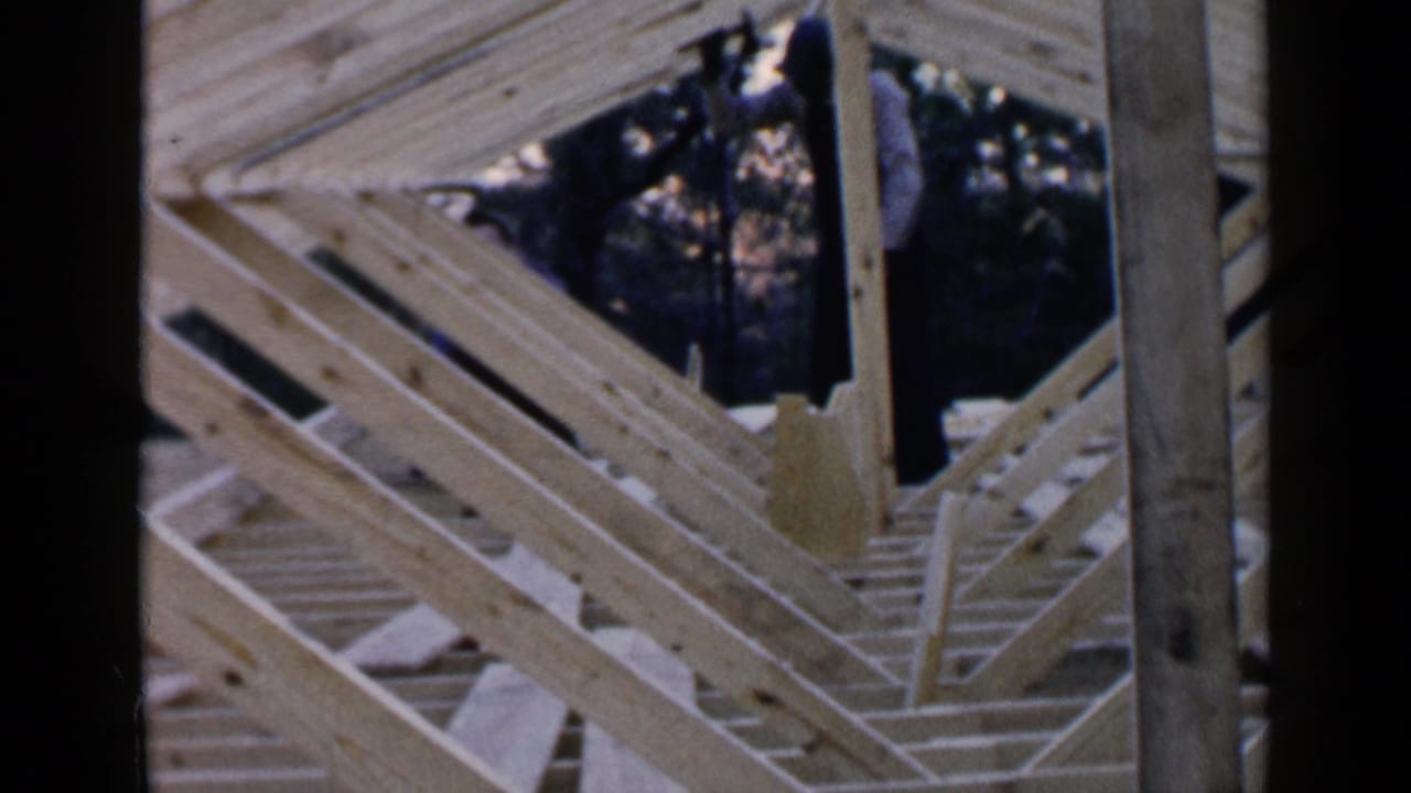 Man in overalls stands among pale wooden roof rafters with a hammer nearby.