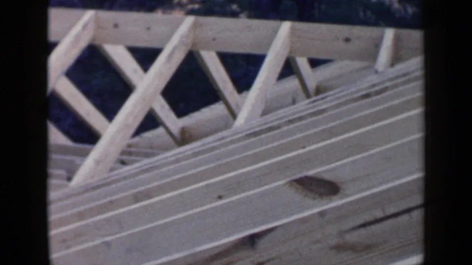 Close 8mm view of pale rafters and diagonal bracing in the unfinished roof frame, with trees beyond.