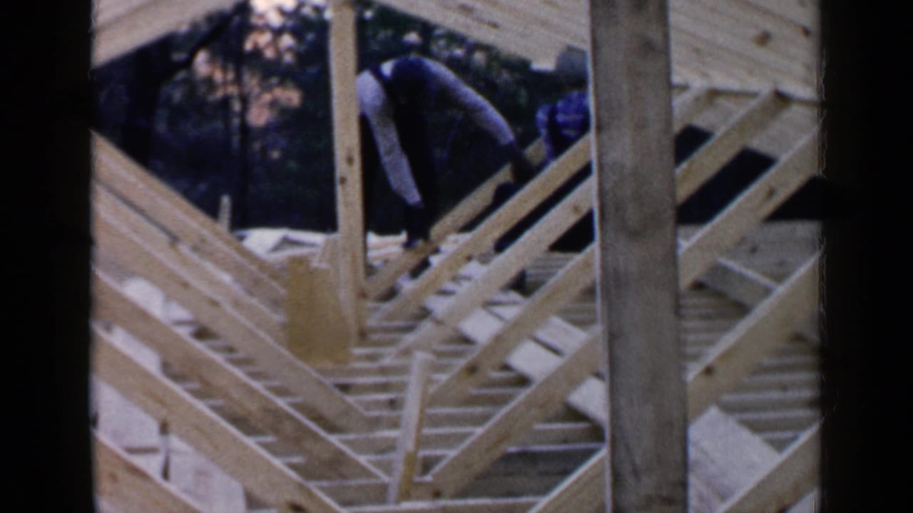 Two men work among unfinished roof rafters on a wooden house frame.