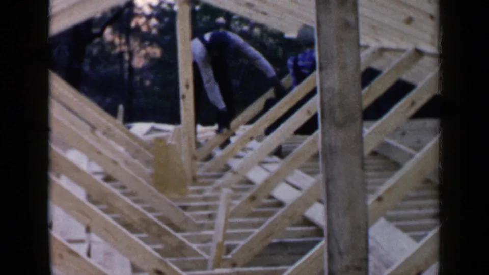 1957 Birmingham home-movie frame showing two men bending over unfinished rafters and vertical posts on a house roof structure.