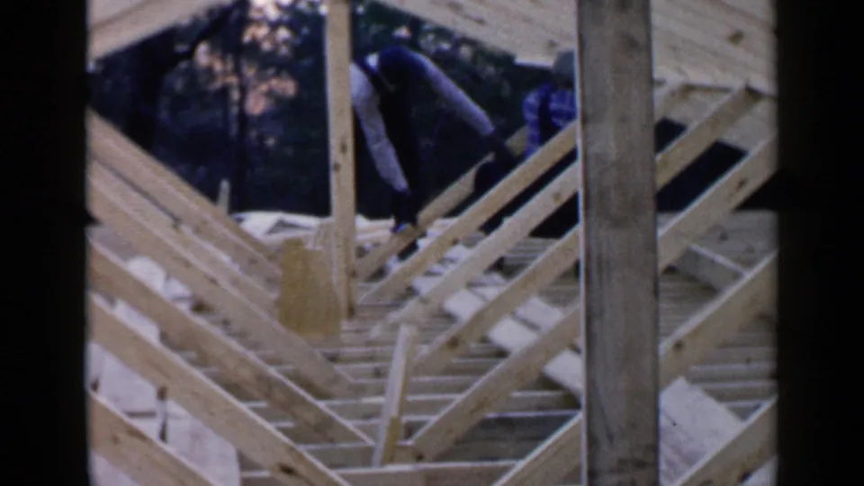 Two men appear on the roof frame of a partially built Birmingham house, surrounded by rafters, posts, and diagonal bracing.