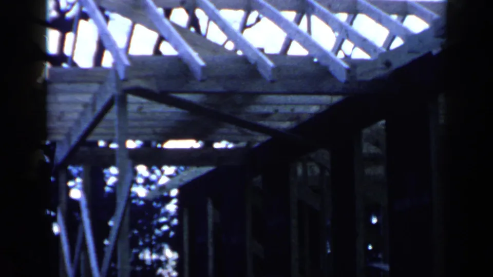Low-angle 1957 Birmingham view beneath a partially framed house roof, showing rafters, posts, braces, and daylight through the structure.