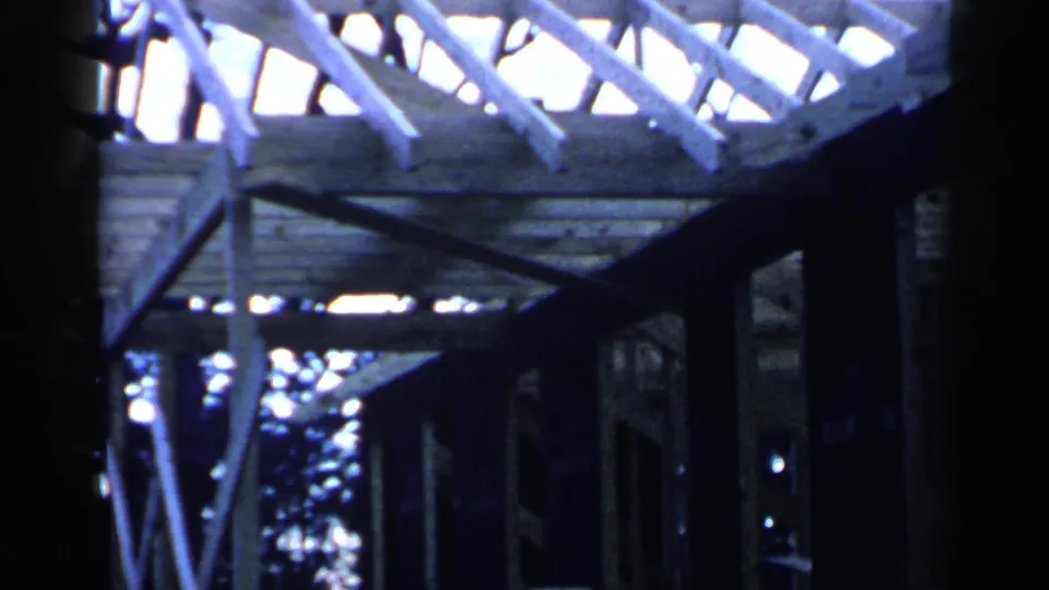 Archival home-movie frame from below the house structure, showing wooden supports and roof framing in strong daylight.
