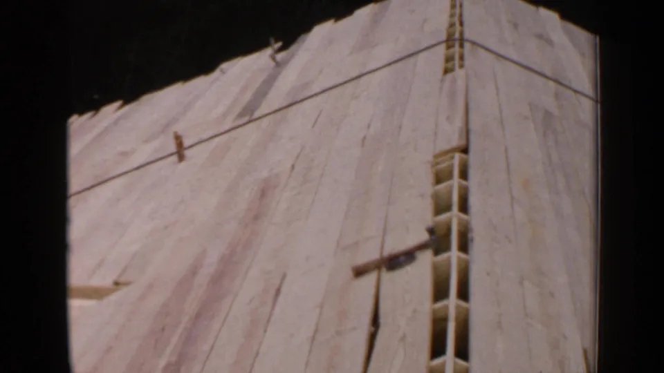 1957 Birmingham home-movie still of sloped wooden sheathing, showing a narrow vertical opening, raw boards, and shadowed framing above.