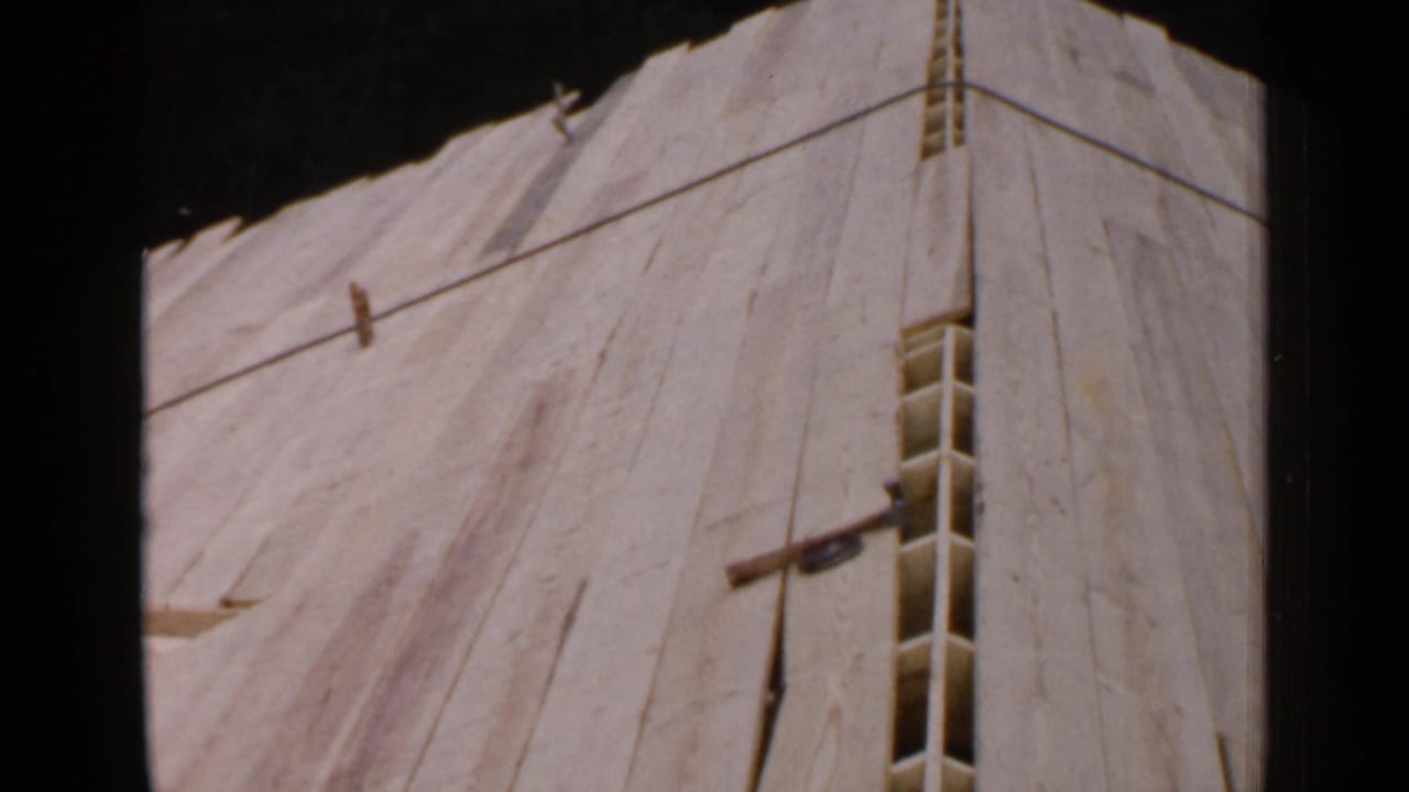 1957 Birmingham 8mm frame showing an unfinished sloped wooden roof structure with plywood panels, narrow framing gaps, and trees in the background.