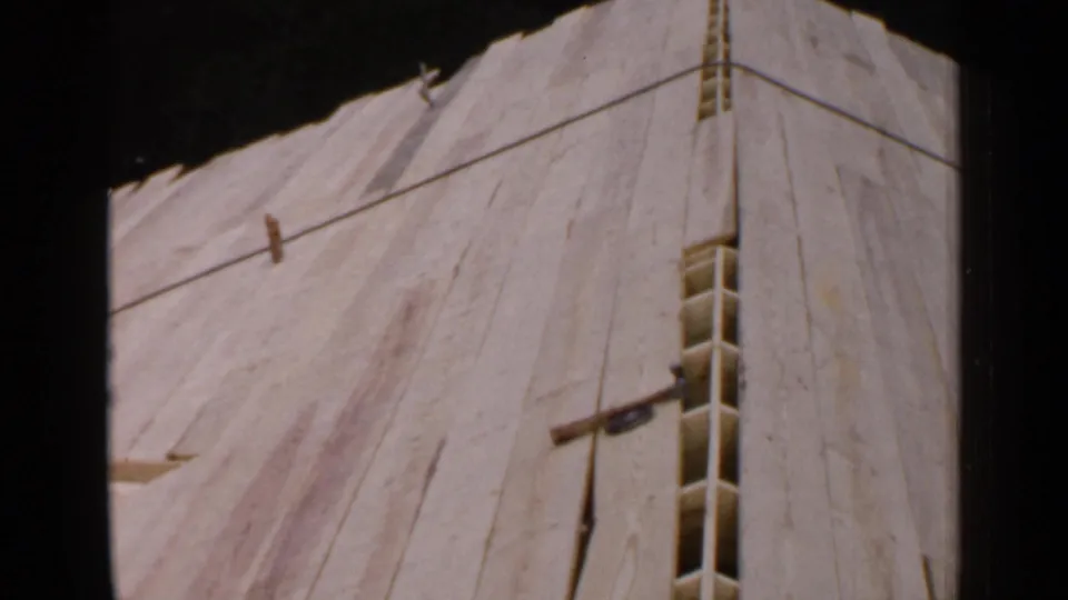 1957 Birmingham 8mm frame showing an unfinished sloped wooden roof structure with plywood panels, narrow framing gaps, and trees in the background.