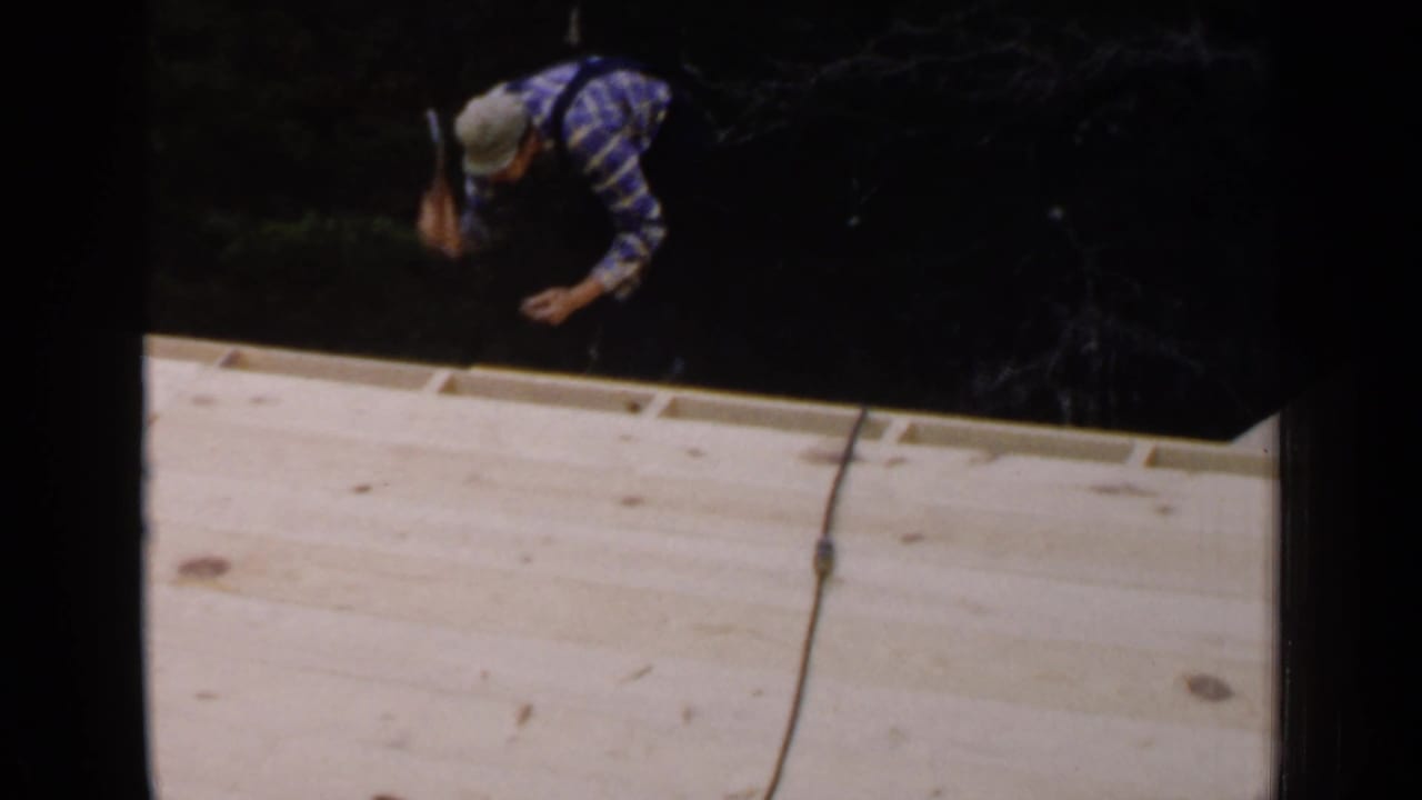 Man in a plaid shirt and cap works along the edge of an unfinished wooden roof.