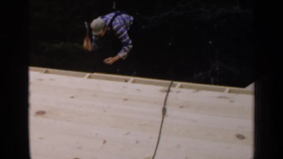 An adult man in a plaid shirt and cap leans over the edge of an unfinished wooden roof at the Birmingham construction site.