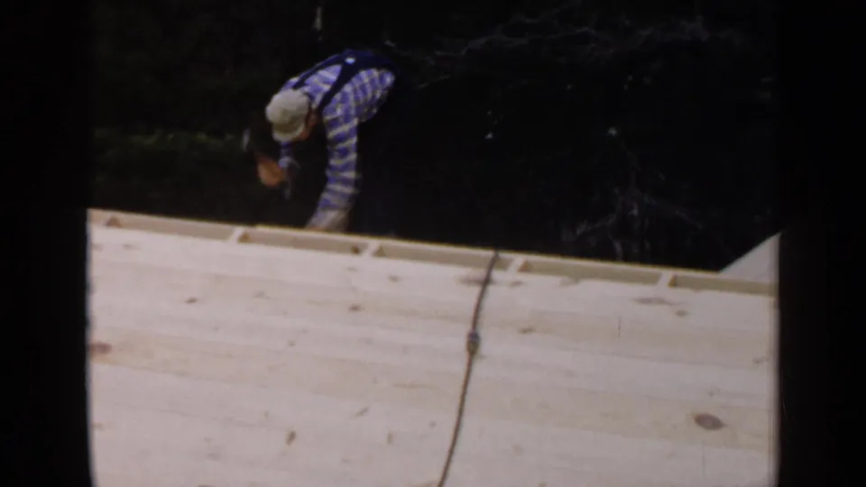 An adult worker in a plaid shirt bends over the unfinished wooden roof edge, with a dark line or cord hanging across the foreground.
