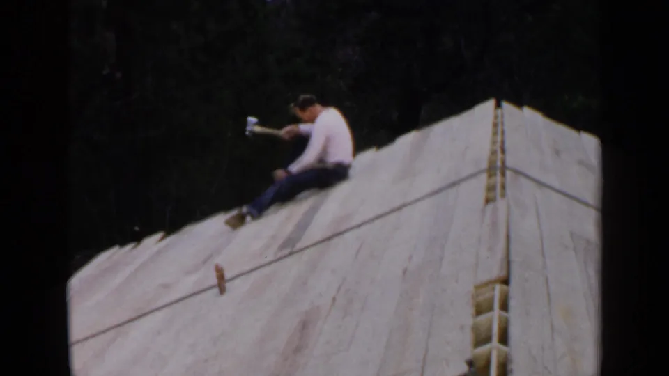 An adult man in a light shirt sits on the sloped unfinished roof in Birmingham, working with a hammer near exposed wooden framing.
