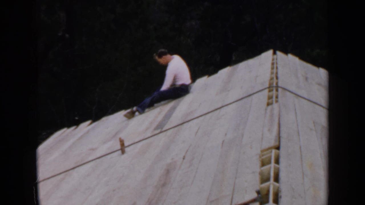 In 1957 Birmingham-area 8mm footage, a man sits on a sloped wooden structure covered with white sheeting against dark trees.