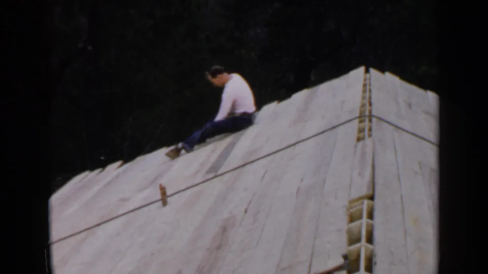In 1957 Birmingham-area 8mm footage, a man sits on a sloped wooden structure covered with white sheeting against dark trees.