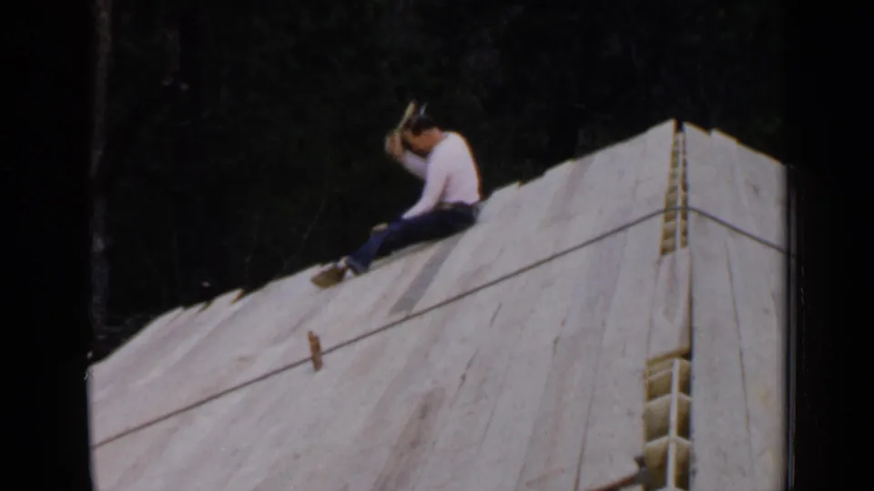 A man in a white shirt raises a hammer while sitting on a sloped white-covered wooden structure in 1957 Birmingham home-movie footage.
