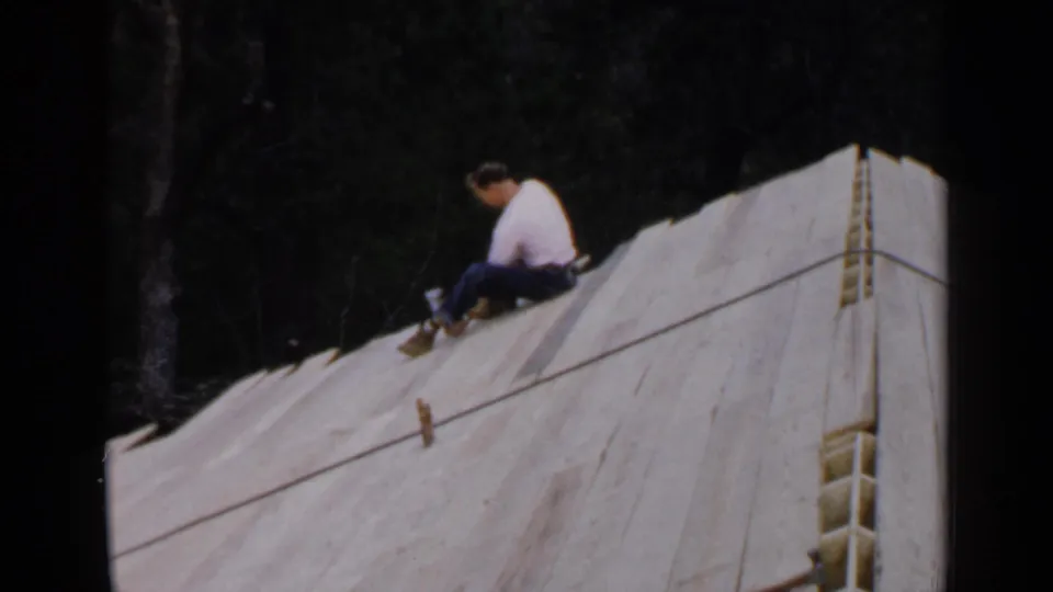 The man remains seated on the sloped white-covered roof structure, looking down toward the surface as trees fill the background.