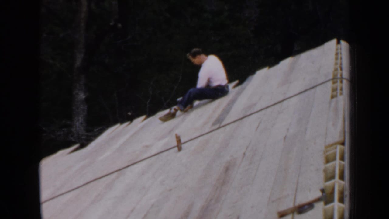 In Birmingham, Alabama home-movie footage from 1957, a man in a white shirt sits on unfinished roof boards near a wooded background.