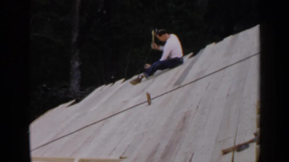 A 1957 Birmingham roof construction view shows a man seated on unfinished wooden sheathing, appearing to work with a hand tool.