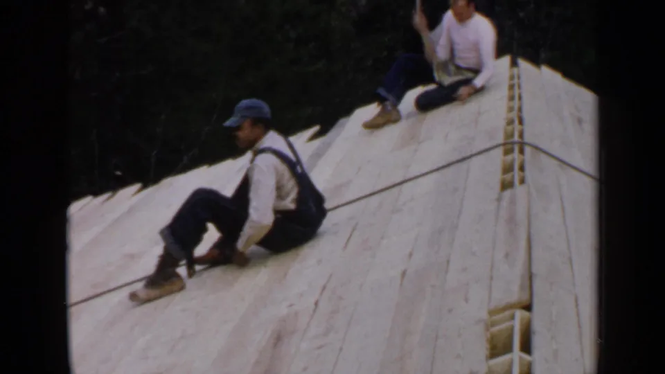Two men are visible on a sloped wooden roof in 1957 Birmingham footage, one in overalls and a cap and another seated higher up.