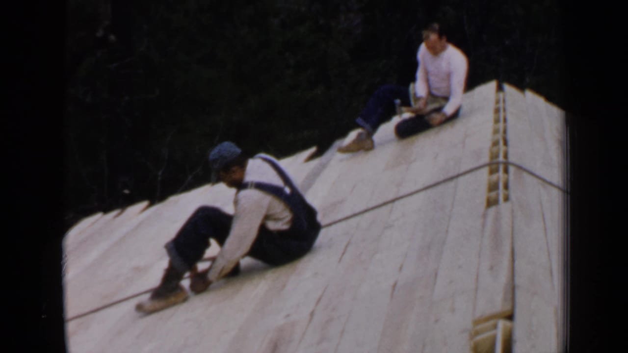 Two men work on a sloped wooden roof, one seated near boards and another farther up with a tool.