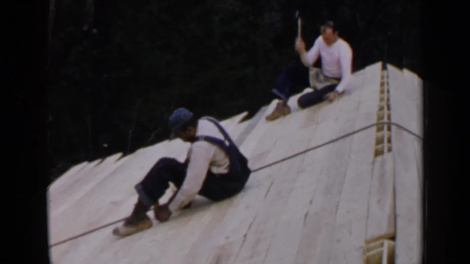 Birmingham, Alabama, 1957 frame showing roof workers on a wooden deck, one lifting a hammer while seated near the ridge.