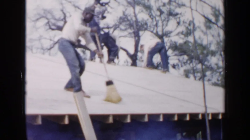 Birmingham, Alabama, 1957 home-movie frame with workers spread across a pale roof surface, including a man sweeping near the edge.