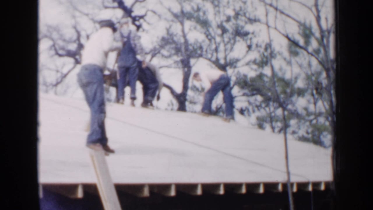 In 1957 Birmingham home-movie footage, several men work on a pale sloped roof with bare trees behind them.