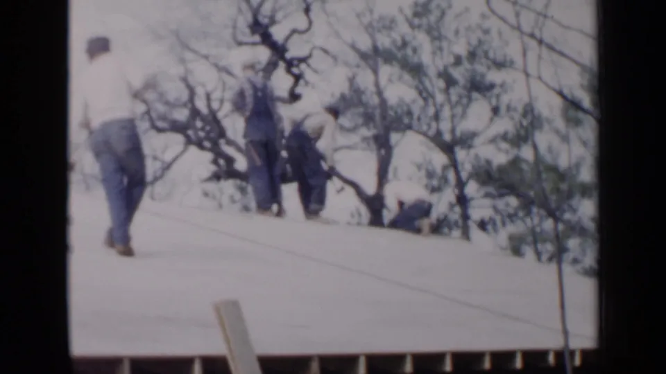 A distant 8mm frame shows men on a sloped roof in Birmingham, with one walking upward and others working near trees.