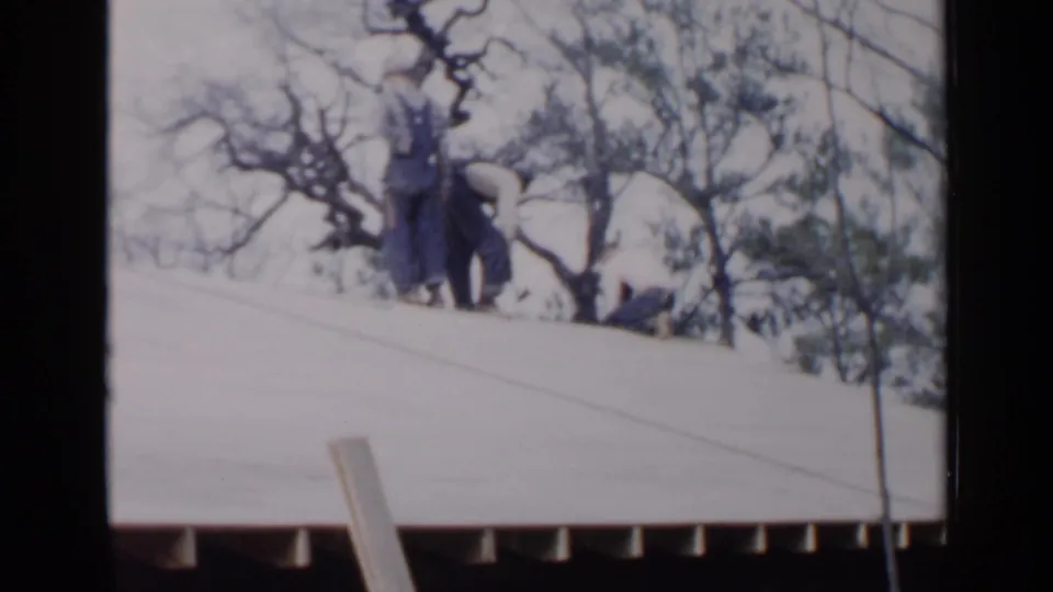 1957 Birmingham home-movie frame showing people on a sloped roof above a house, partly obscured by bare winter trees.