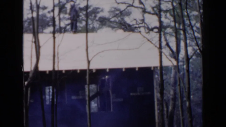 1957 8mm frame of a dark residential exterior and pale roof seen through leafless trees, with bluish haze near the lower frame.