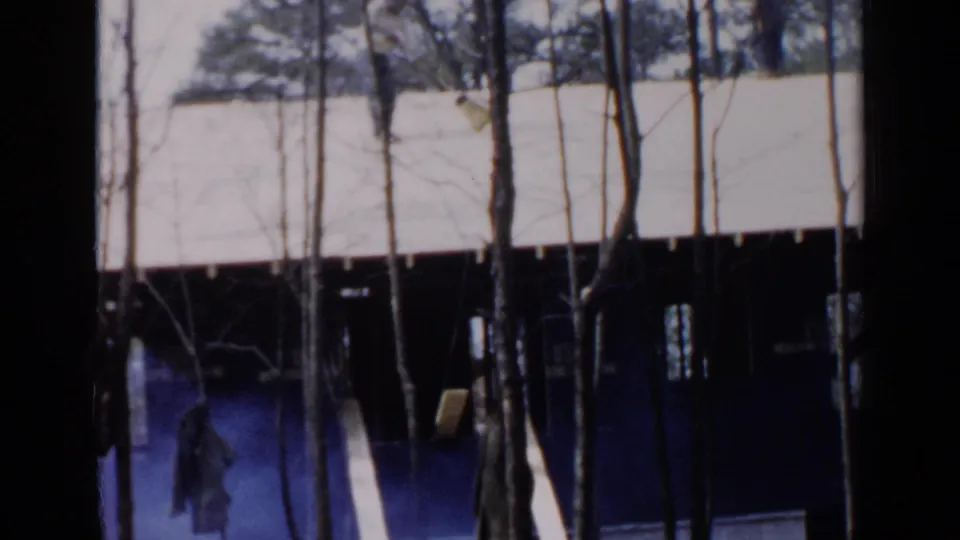 A person stands near the lower edge of a dark house exterior in 1957 Birmingham footage, with bare trees and a pale roof overhead.